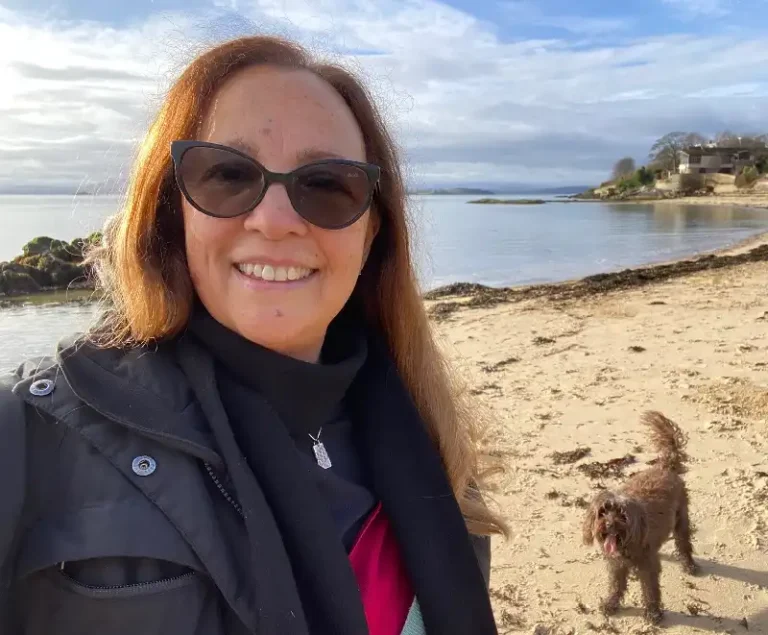 Ginnie Monk, contemporary Scottish jeweller standing on a sandy beach with a curved bay behind her.